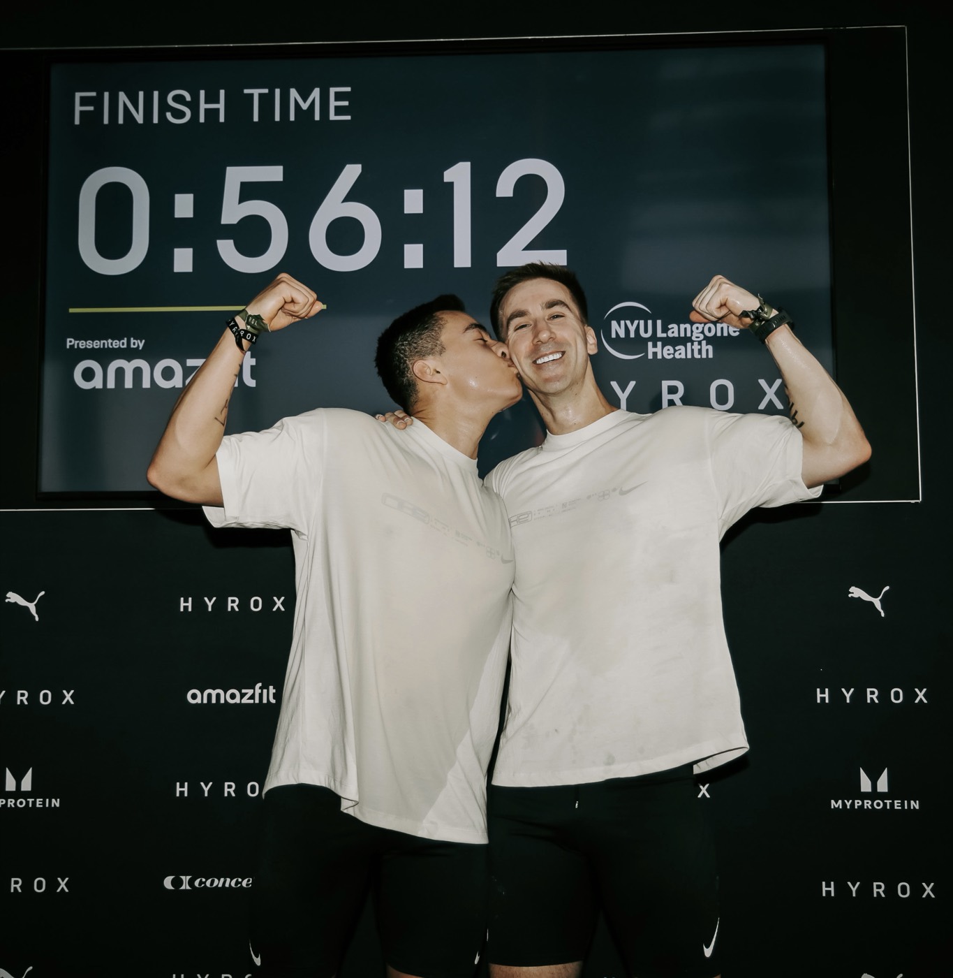 Cesar and Derek flexing in front of the 0:56:12 finish time board at Hyrox NYC