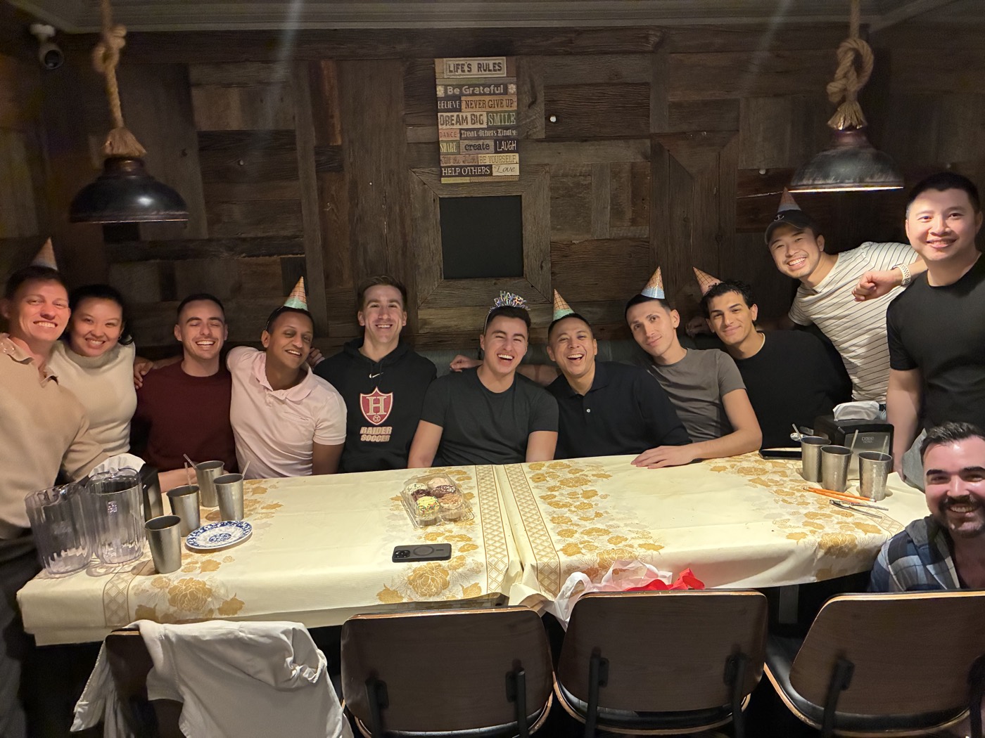 A long birthday dinner table of friends in party hats, everyone laughing